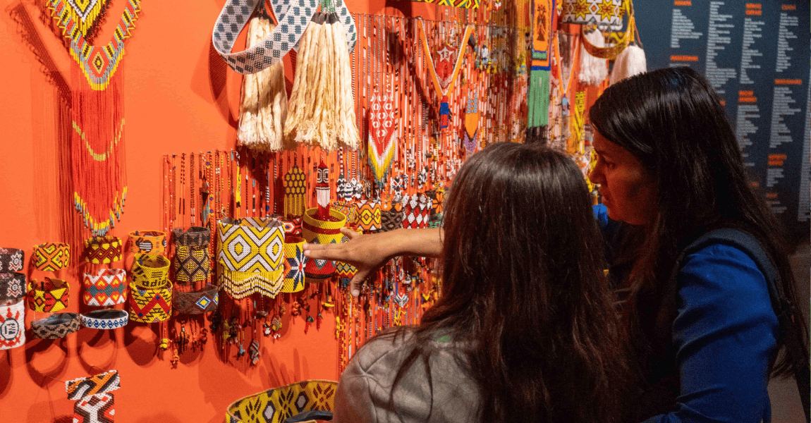 Mulheres observam e apontam para enfeites indígenas expostos em uma parede laranja