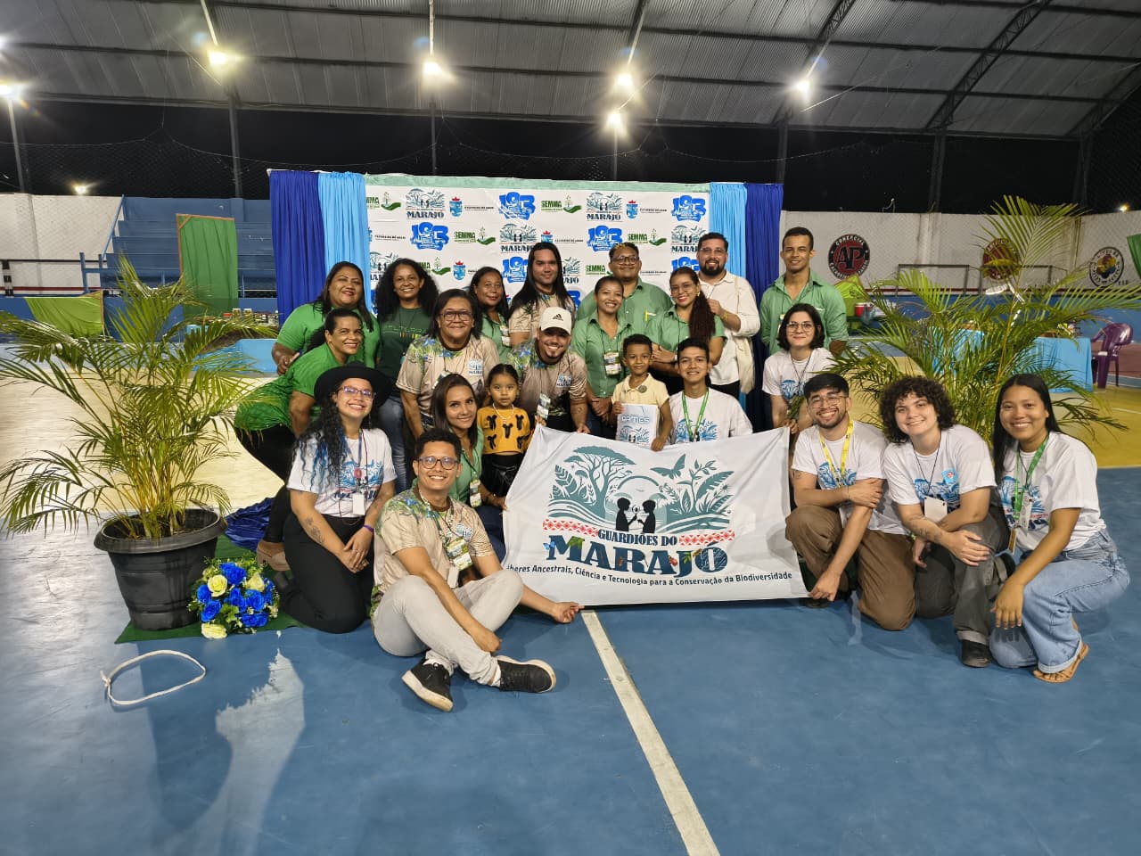 Em um ginásio, a equipe organizadora do evento posa para a foto segurando uma bandeira
