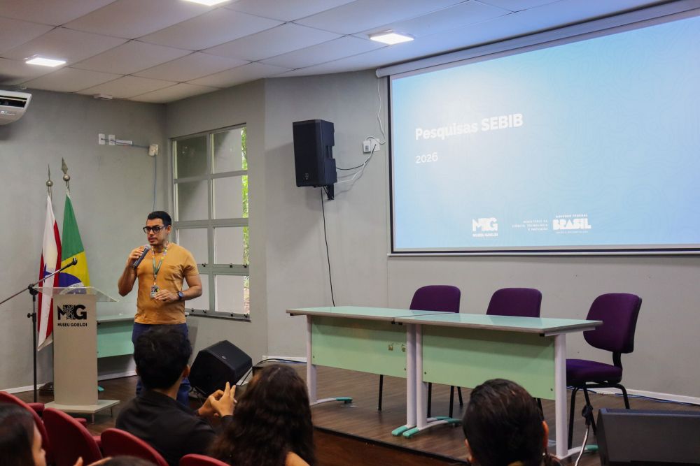 Homem segurando microfone fala a pessoas em auditório