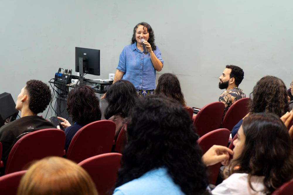 Mulher sorri ao falar a plateia em um ambiente de auditório