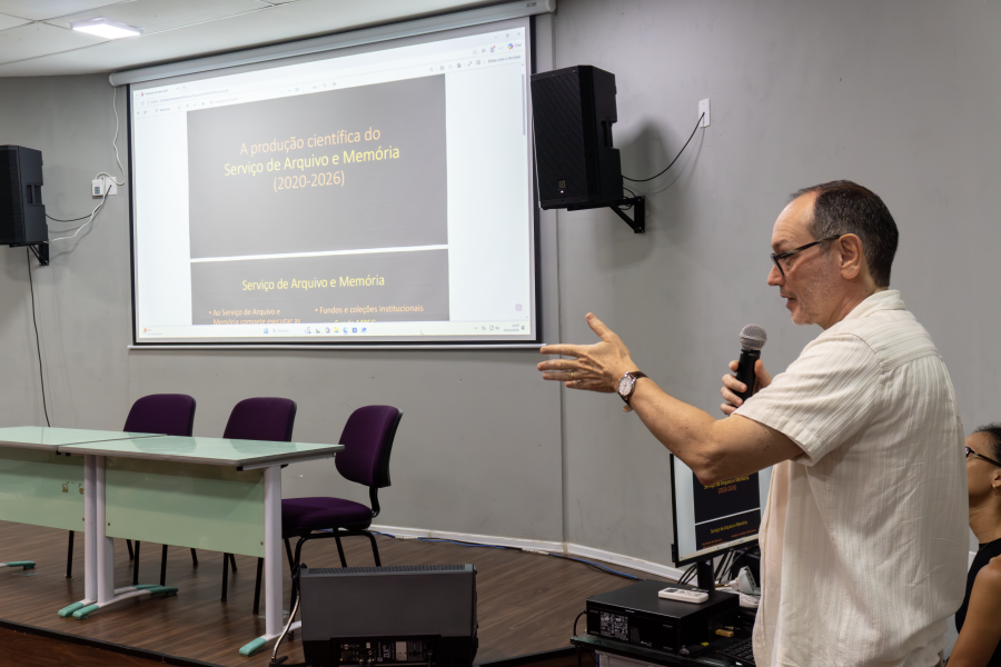 Homem fala a uma plateia apresentando slide