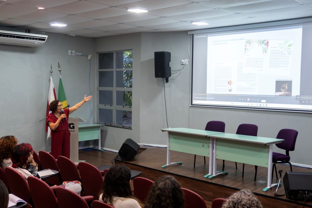 Mulher em pé em auditório fala a plateia apontando para projeção em telão