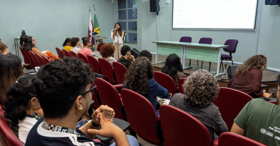 Jovem segurando um microfone fala a uma plateia. No fundo, um texto exibido em um telão