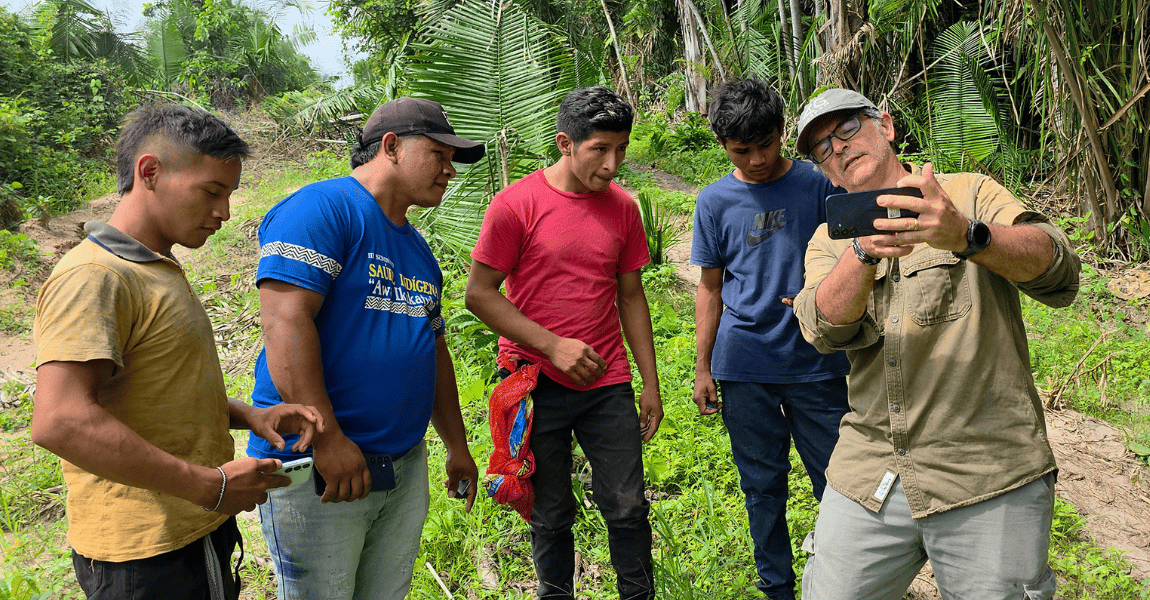 Em uma área de vegetação amazônica, quatro jovens indígenas observam atentamente um homem mais velho, de boné e óculos, que segura um celular com a tela voltada para o grupo, aparentemente mostrando alguma informação ou imagem. Os jovens usam camisetas em tons de amarelo, azul, vermelho e azul-escuro, e alguns também seguram celulares nas mãos. Ao fundo, há palmeiras altas, folhas largas e um caminho de terra cercado pela mata.
