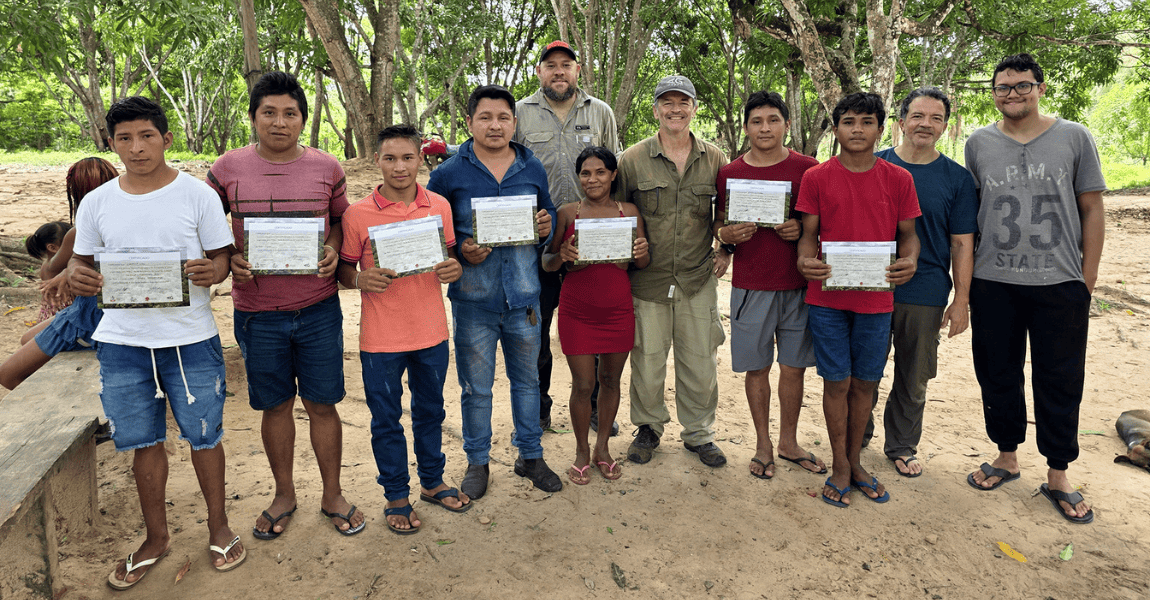 Grupo de onze pessoas posa em pé em uma área aberta cercada por árvores, em ambiente de floresta ama