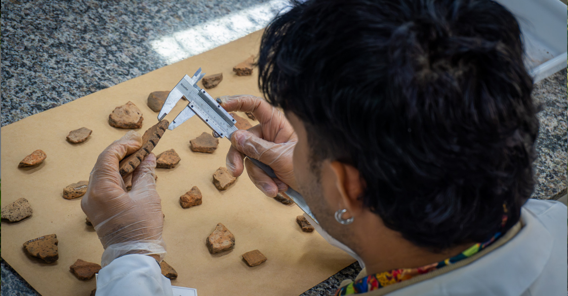 Um estudante está de costas, usando jaleco, segurando uma ferramenta utilizada numa aula de arqueologia, medindo uma peça de cerâmica, usando luvas, diran te de uma mesa com outros fragmentos de cerâmica em cima de uma cartolina