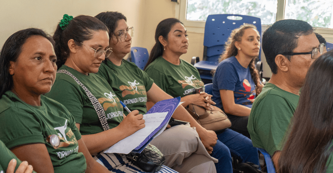 Mulheres vestidas com camisetas verdes da jornada assistem aula. Uma delas escreve num caderno.