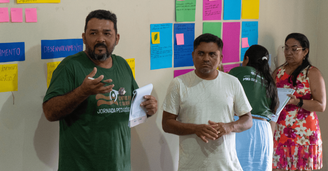 Dois professores estão lado a lado diante de papéis colados na parede em dinâmica de grupo