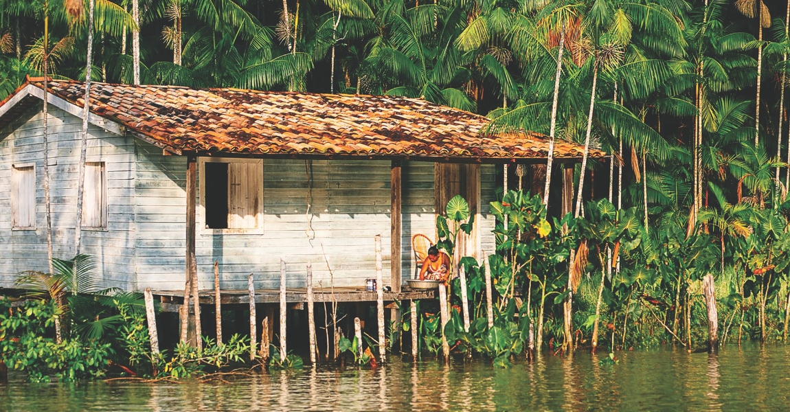 Uma casa simples de madeira sobre estacas fica à beira de um rio, cercada por vegetação tropical densa e altas palmeiras. Na varanda, uma mulher sentada, aparentemente lava ou prepara algo em uma bacia