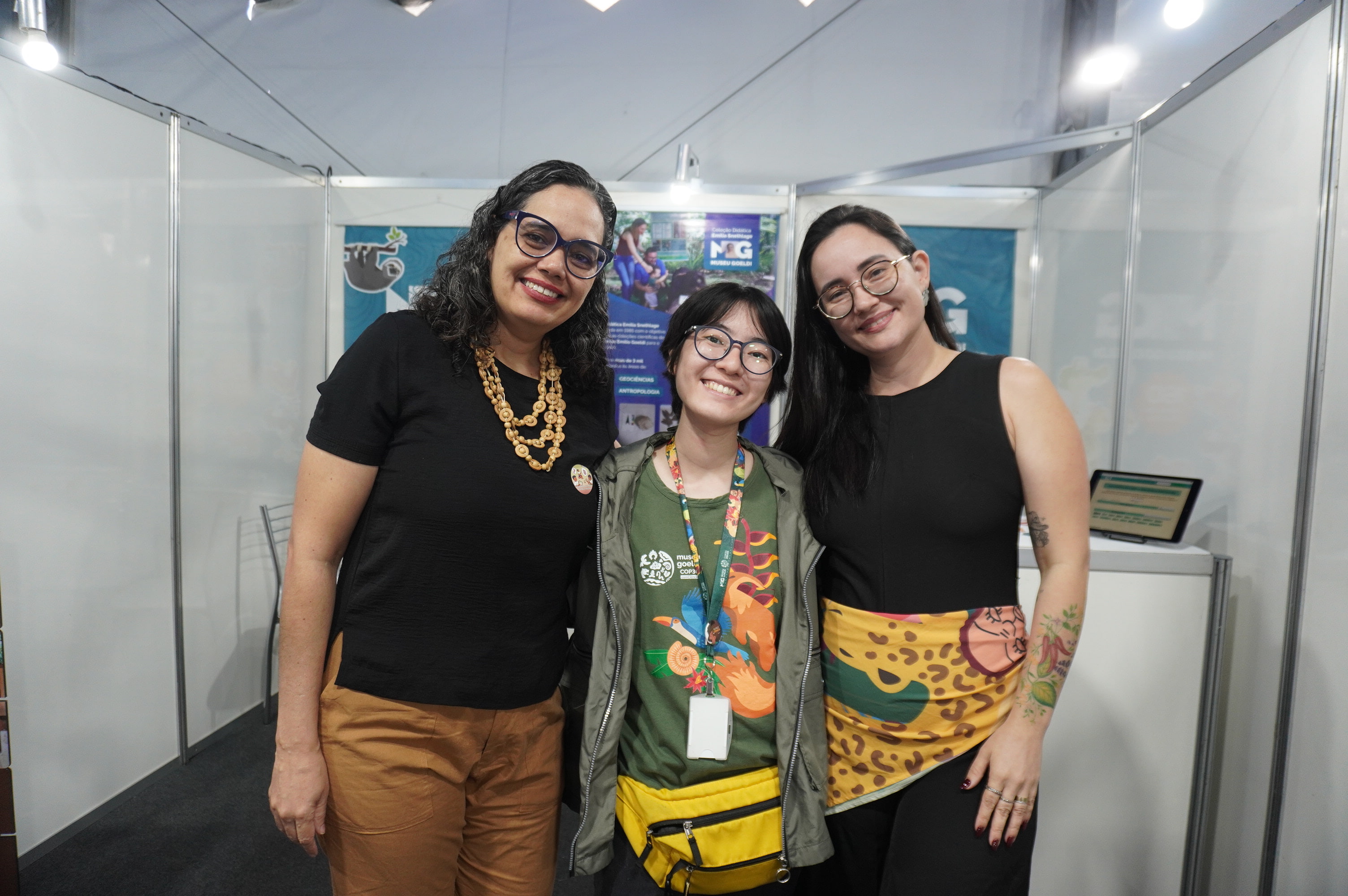 três mulheres posam para foto tendo ao fundo banners coloridos