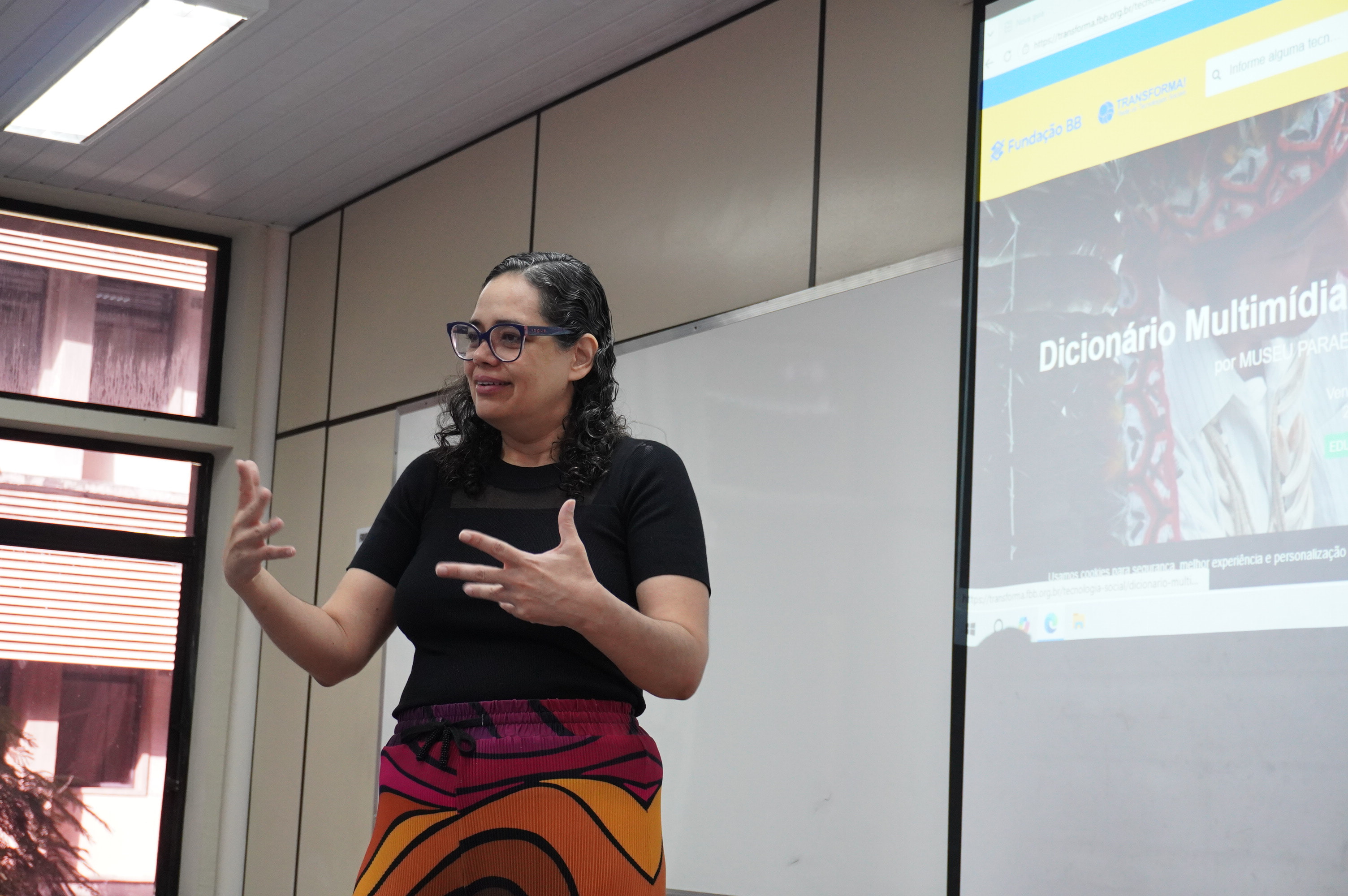 mulher gesticula tendo ao lado um telão com a temática de tecnologia social