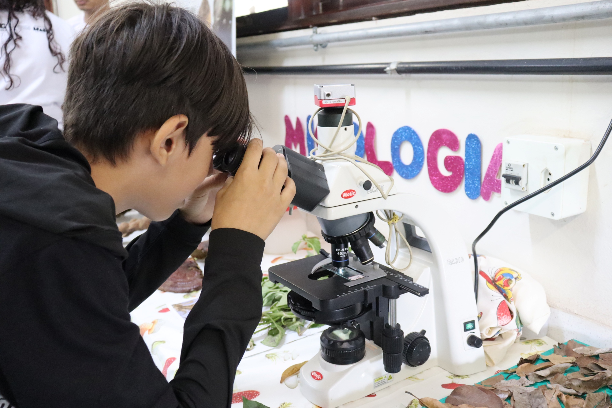 Menino observa uma amostra em um microscópio sobre uma mesa com folhas e materiais de pesquisa