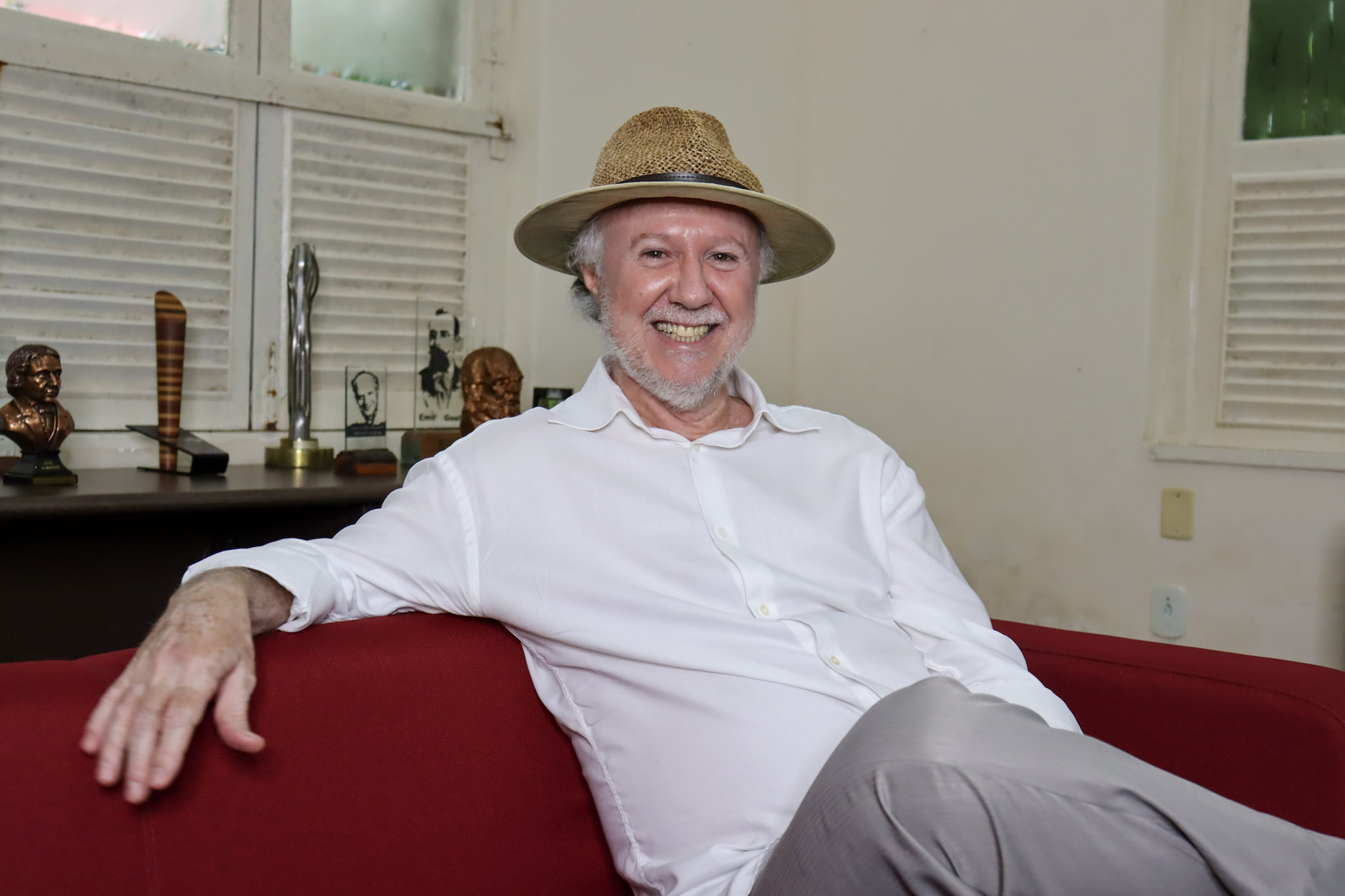 Um homem branco está sentado num sofá vermelho usando chapéu de cor palha e camisa branca