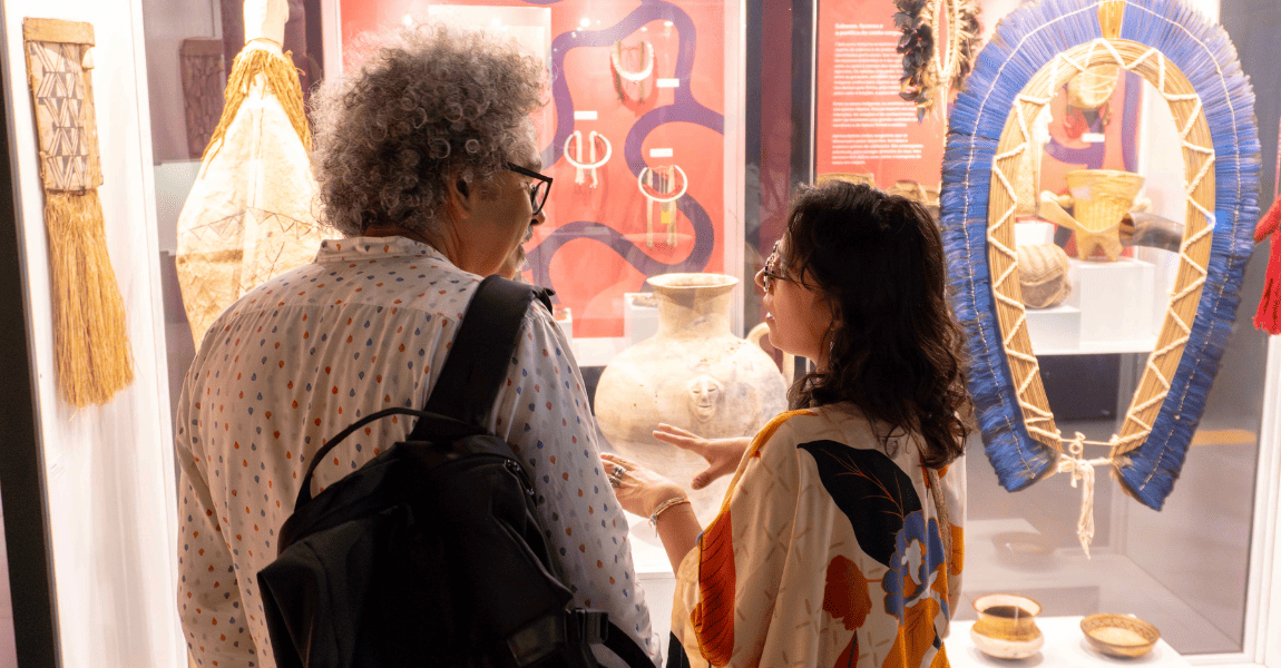 Um homem escuta uma mulher diante de uma vitrine com cocar indígena em exposição