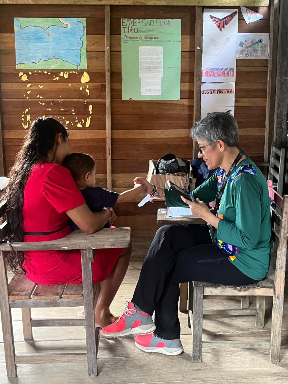 Em uma sala de madeira, uma médica atende uma mulher e uma criança sentadas em uma cadeira