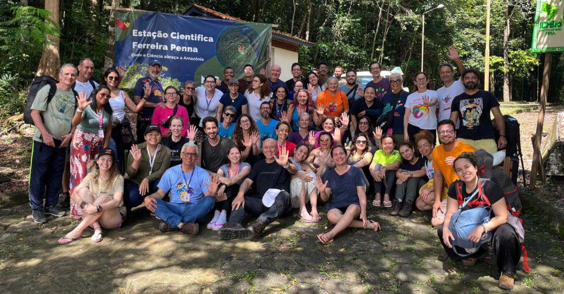 Grupo de voluntários da caravana da saúde posa sorrindo em frente à Estação Científica Ferreira Penna, cercados pela floresta amazônica, acenando para a câmera em um momento de confraternização.