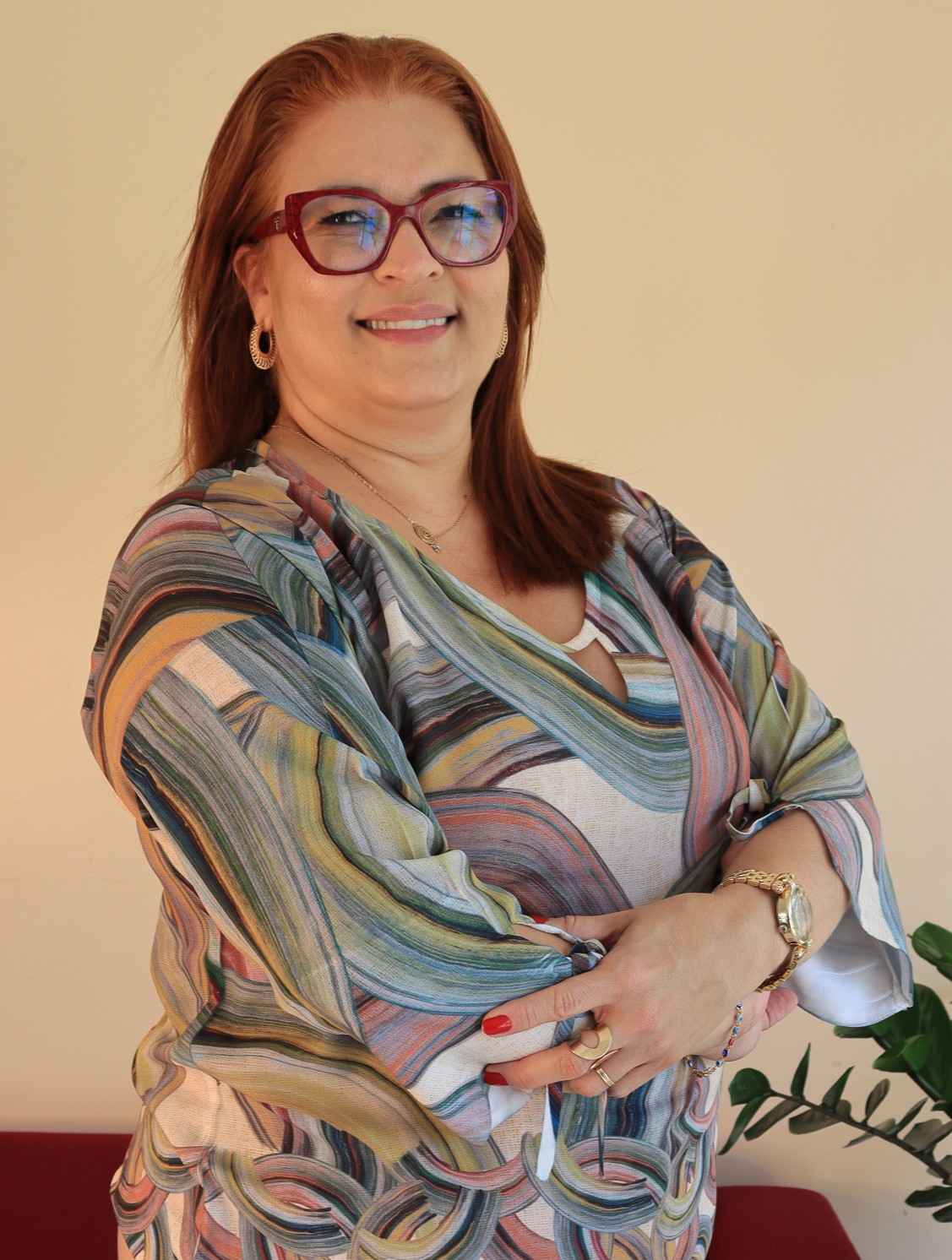 Uma mulher branca de óculos posa para foto de braços cruzados. Ela usa blusa colorida com calça bege