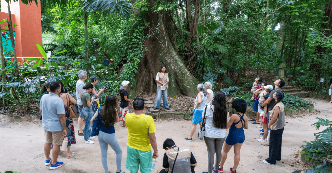 Um grupo de visitantes no parque zoobotânico estão ao redor de uma árvore alta ouvindo a guia fala