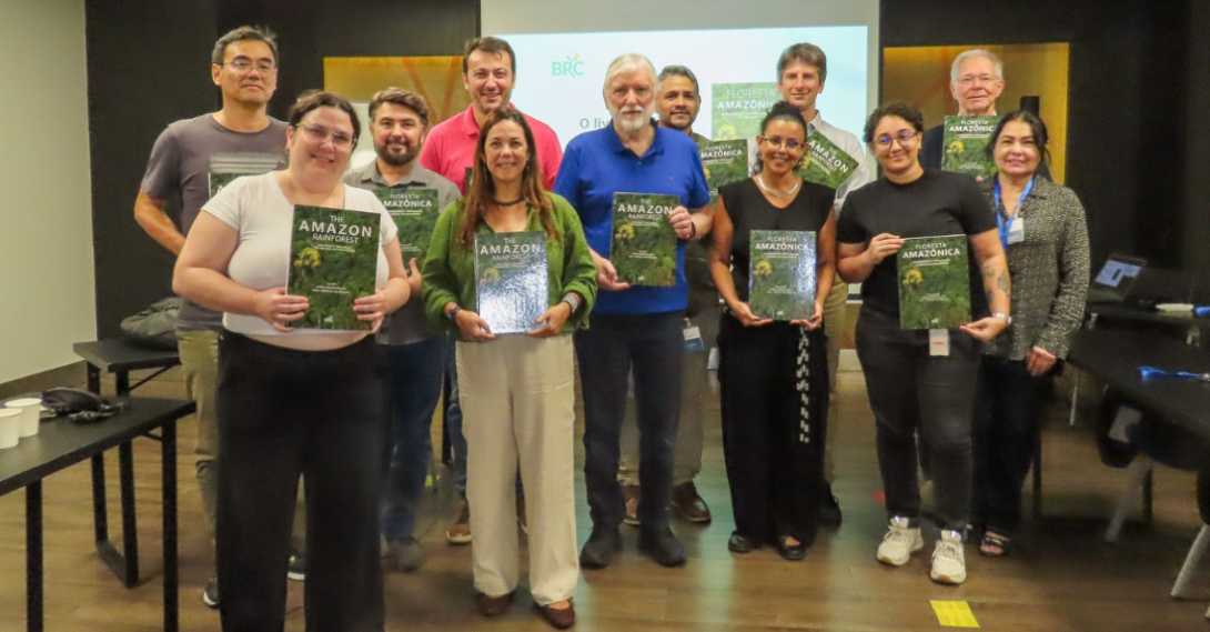 Pesqusiadores homens e mulheres seguram um livro de capa verde com imagem da Amazônia numa sala de reunião