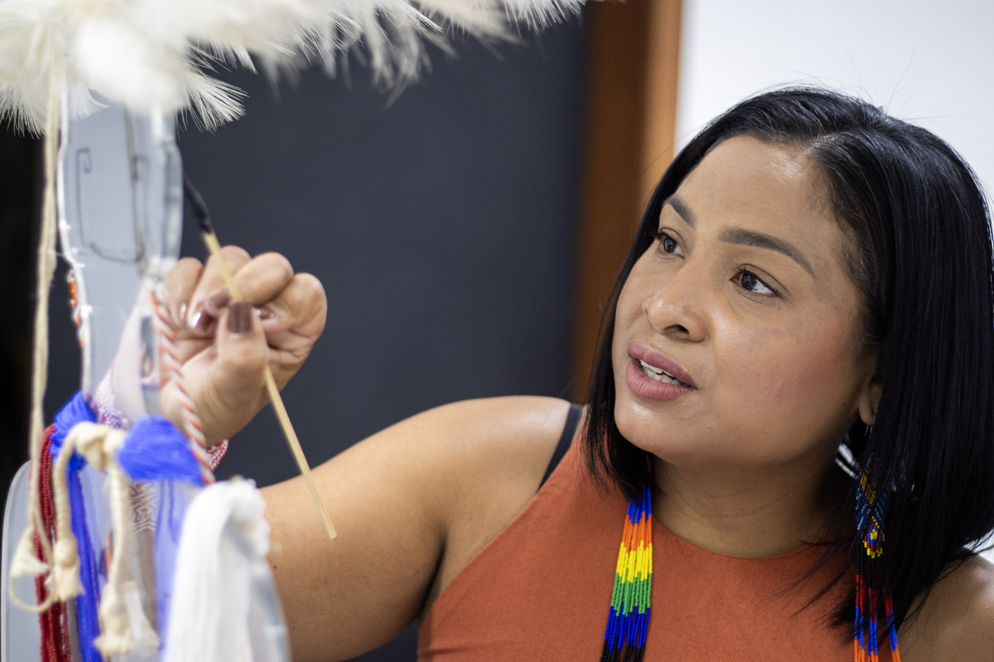 Uma indígena de blusa laranja, brincos coloridos e cabelos pretos pinta um objeto transparente