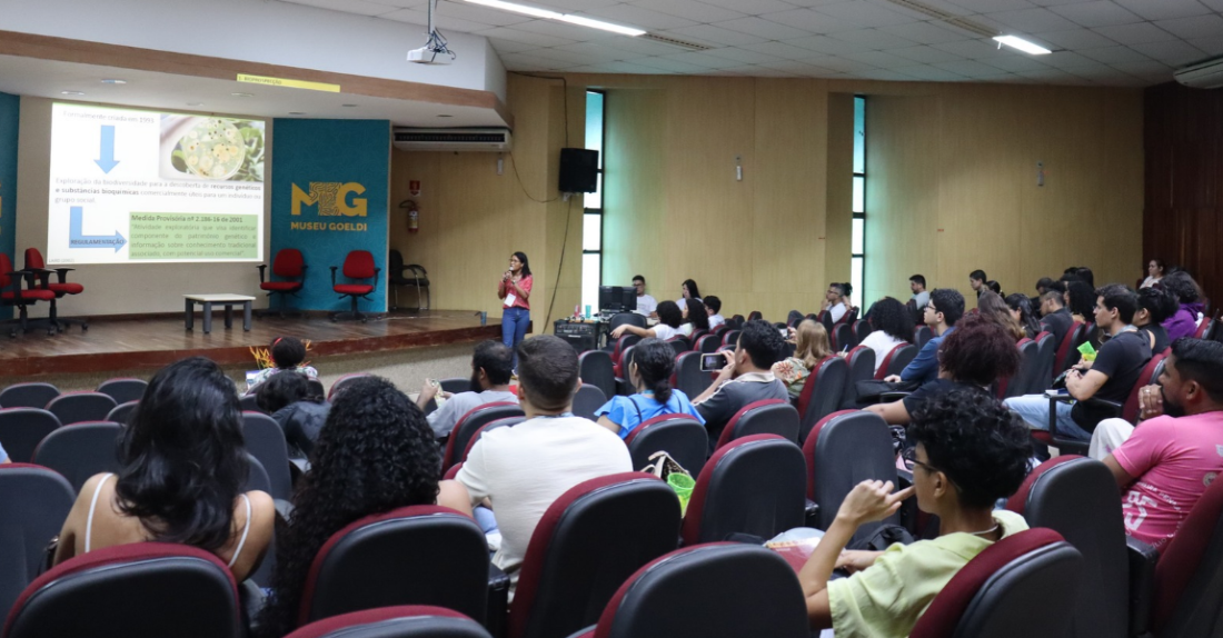 Estudante fala ao microfone para plateia tendo ao fundo uma apresentação de slides
