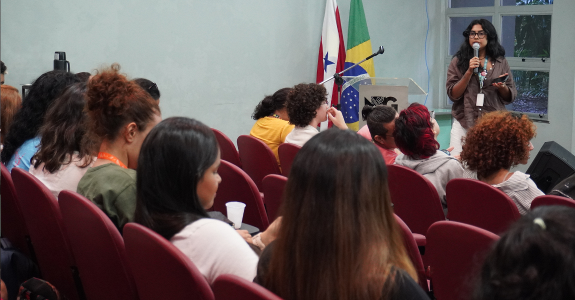 Uma jovem de cabelos pretos fala ao microfone para uma platéia de estudantes