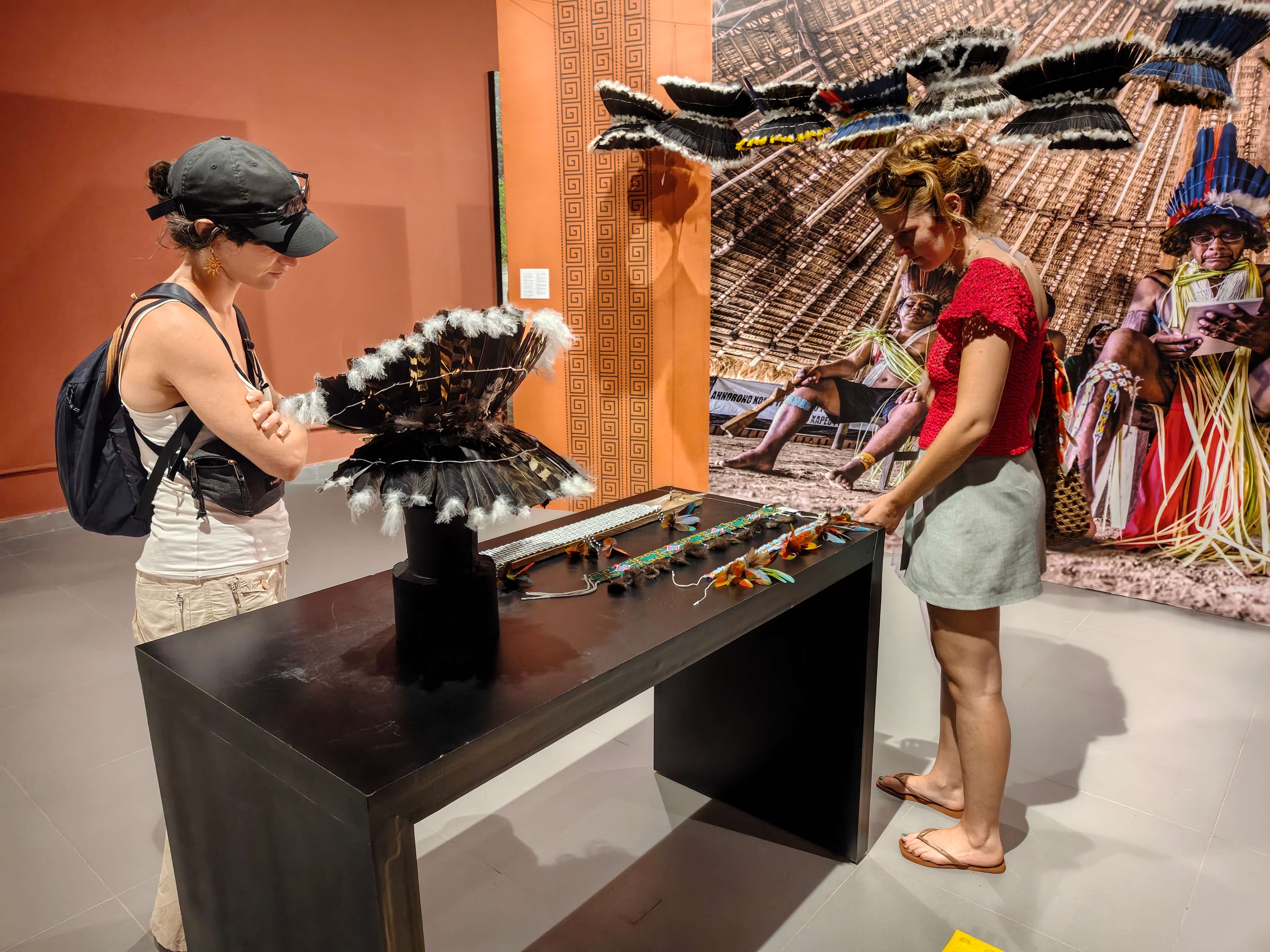 Duas turistas usando roupas leves estão de cabeça baixa olhando uma vitrine na exposição