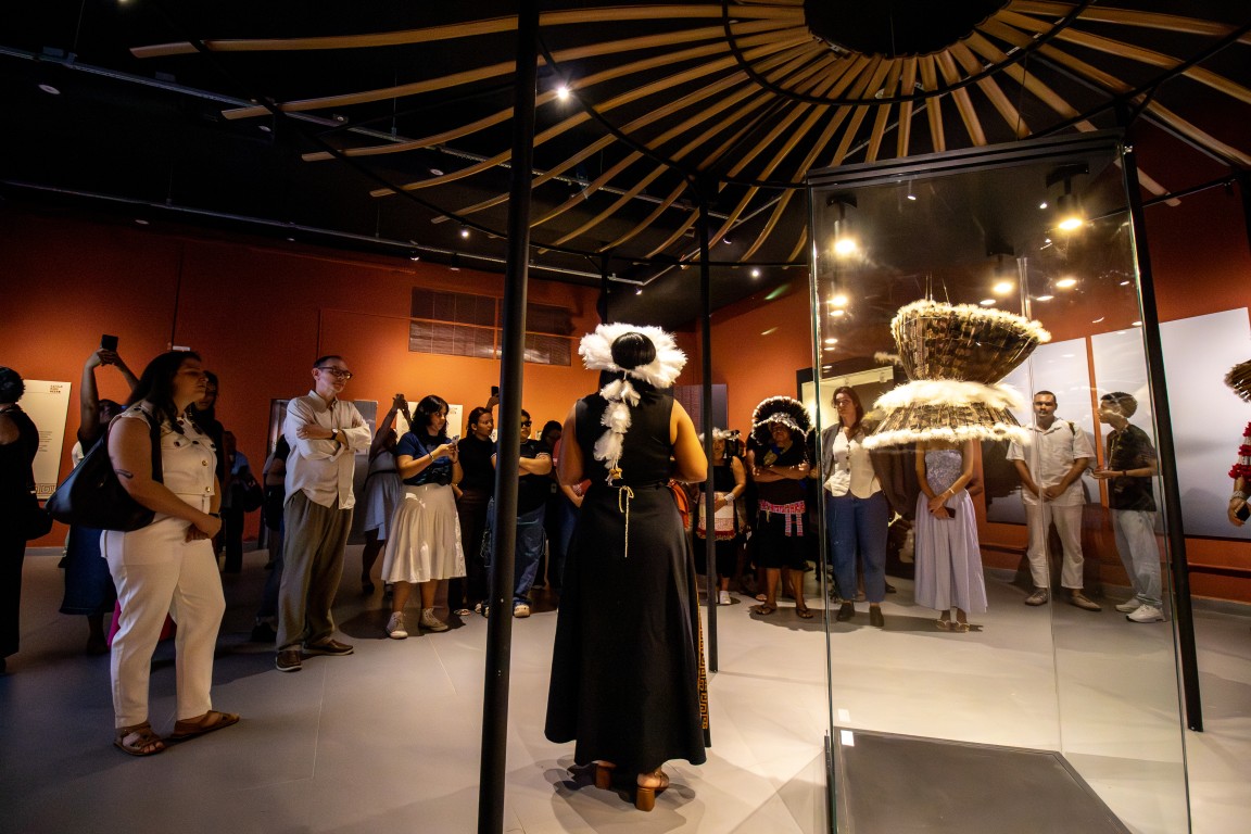 Um grupo de pessoas observam uma mulher indígena falando no centro de uma exposição