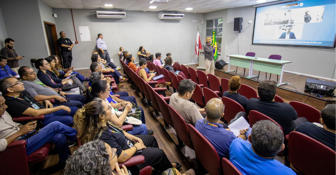 Homem branco usando chapéu fala ao microfone para uma plateia de auditório
