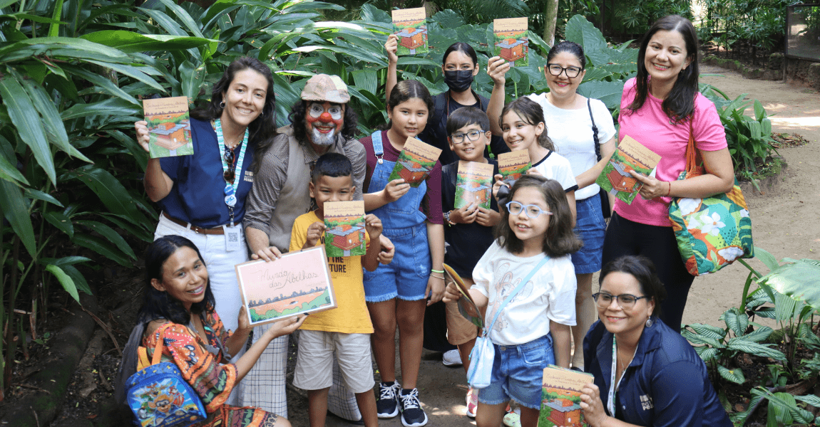 Um grupo de crianças com algumas mulheres adultas e um palhaço posa para foto segurando uma cartilha sobre abelhas numa área verde do parque zoobotânico