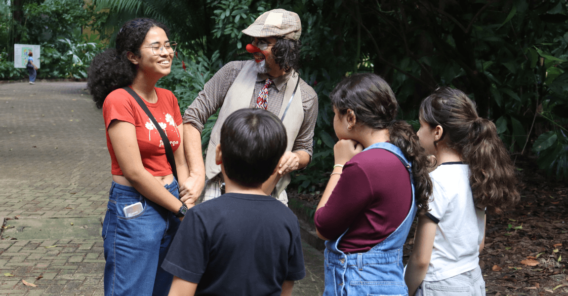 Um palhaço conversa sorridente com quatro crianças que pretam atenção. Eles estão numa área verde.
