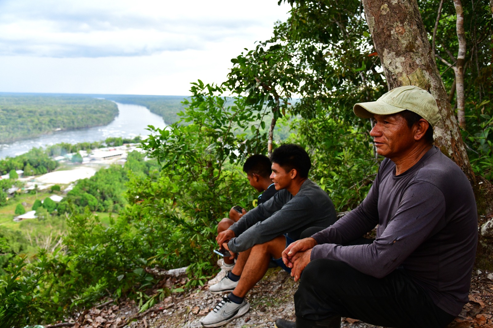 três homens indígenas sentados no alto de uma serra observando um rio ao longe e abaixo