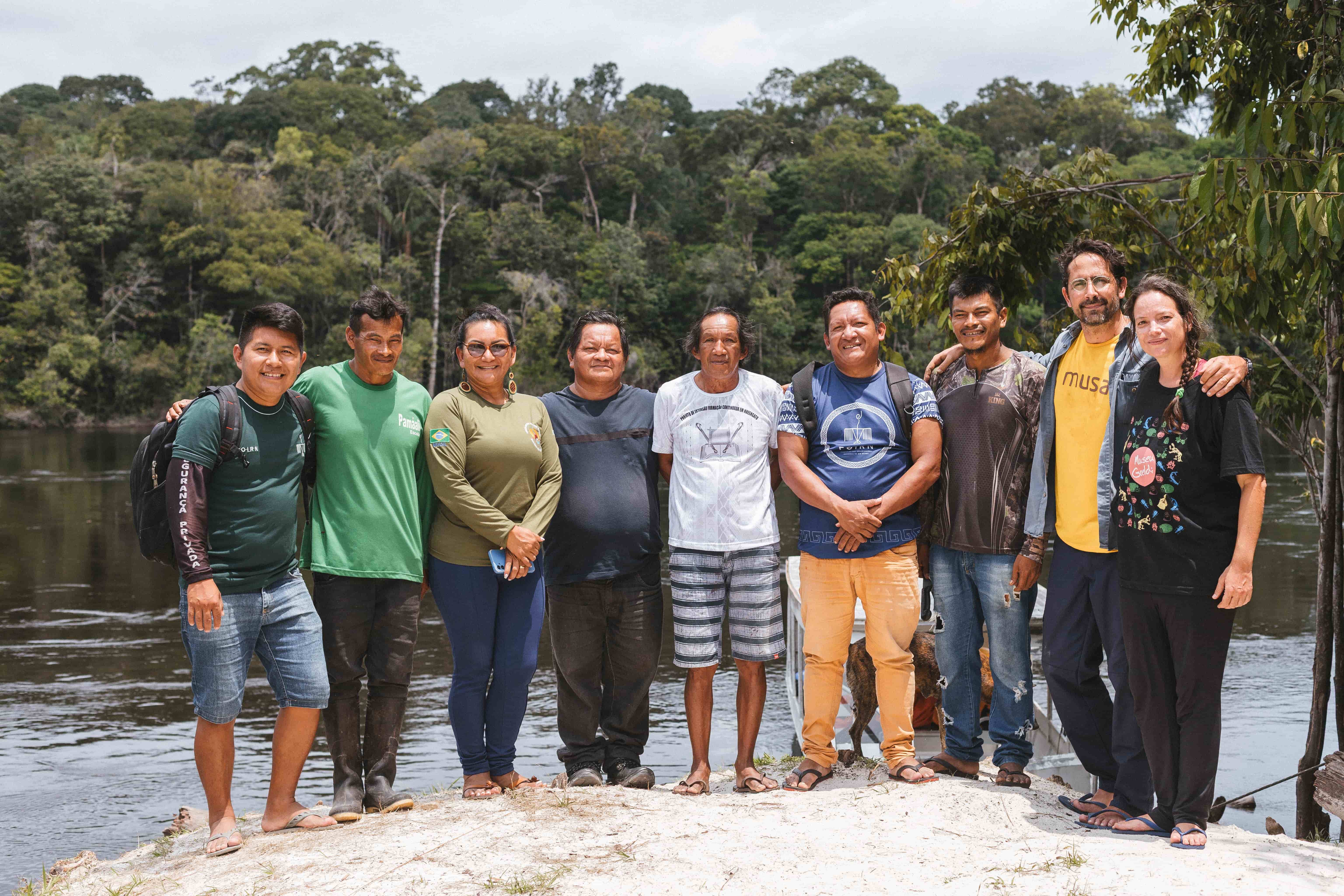 Grupo de nove pessoas, homes e mulheres, alguns indígenas, posam para foto á beira de rio