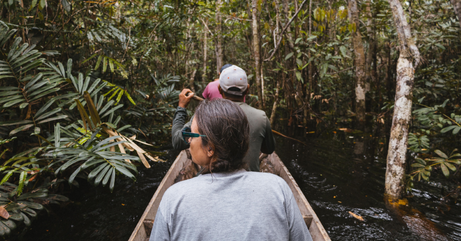 Três pessoas em canoa navegam em rio escuro ladeado de floresta