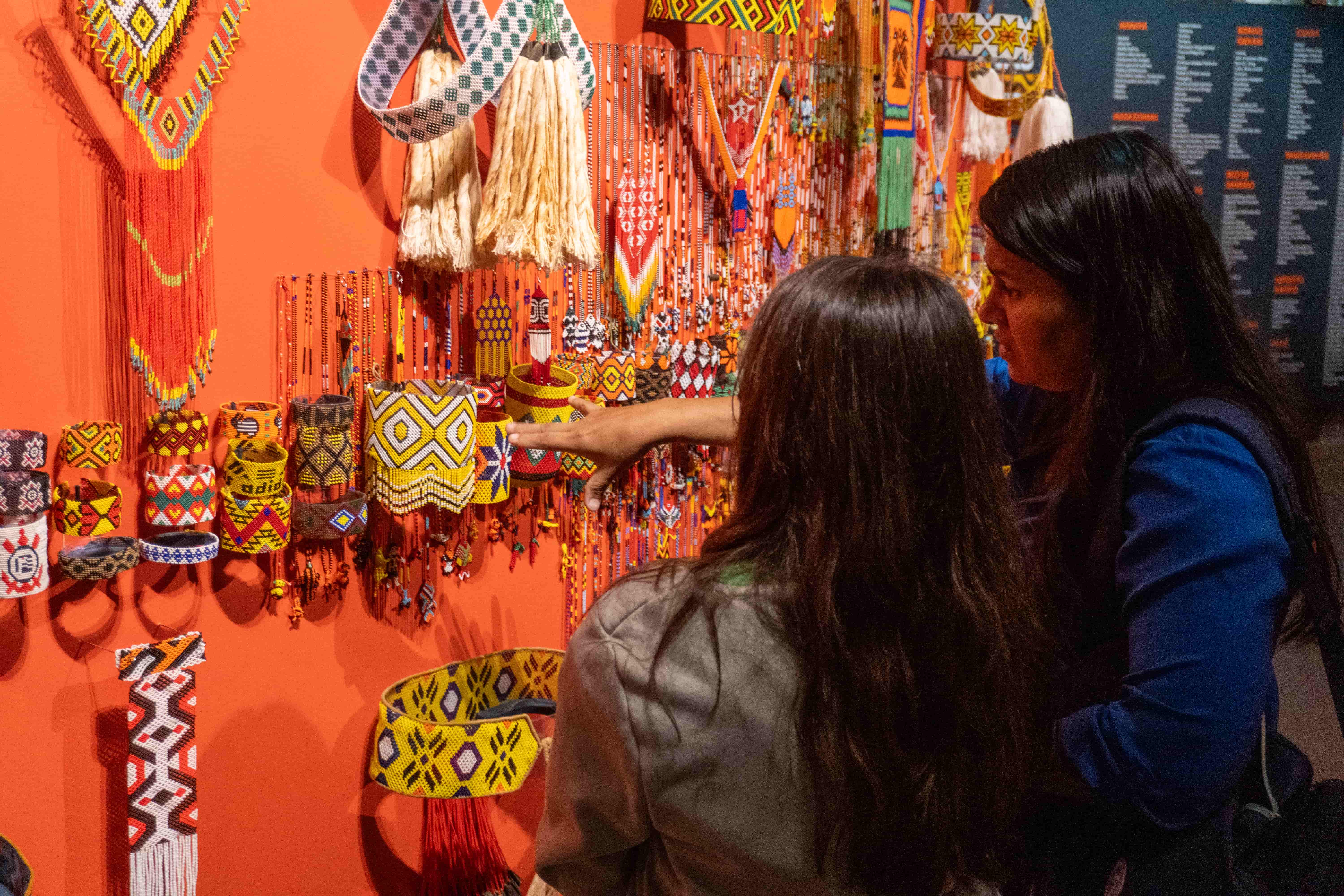 Duas mulheres observam enfeites indígenas expostos em parede e uma delas aponta para uma peça