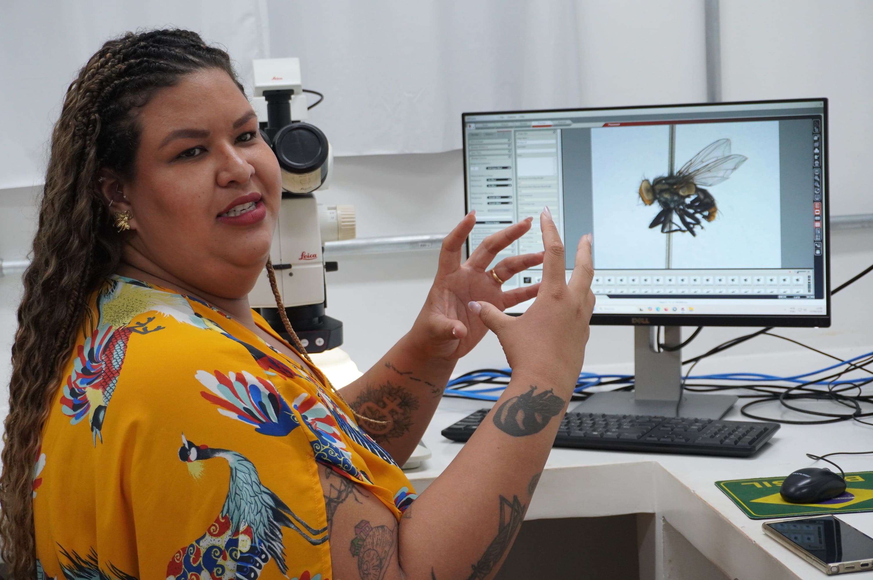 Fotografia em ambiente de laboratório. Em primeiro plano, à esquerda, uma mulher negra de cabelos longos, trançados na parte superior e cacheados nas pontas, sorri enquanto gesticula com as duas mãos abertas. Ela veste uma blusa amarela estampada com aves coloridas e possui diversas tatuagens nos braços. Ao fundo, em uma bancada branca, há um microscópio estereoscópico branco da marca Leica e um monitor de computador ligado. Na tela do computador, é exibida a imagem em alta resolução de uma mosca fixada em uma alfinete entomológico, acompanhada de um software de análise de imagem.