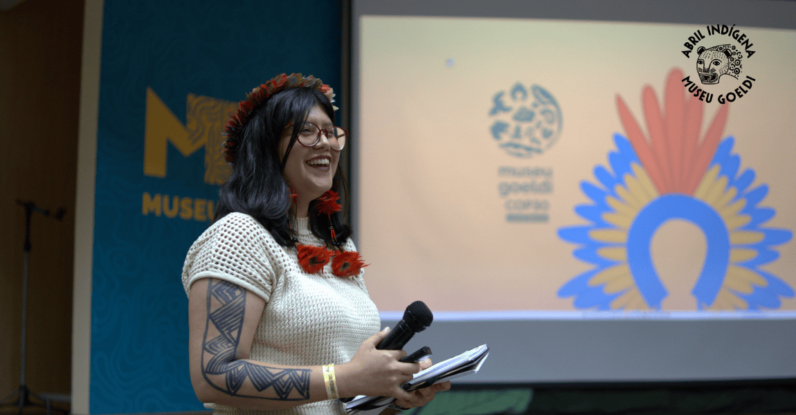 Mulher jovem usando óculos e com adornos indígenas, sorrindo e segurando um microfone