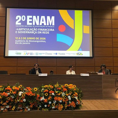 **Texto alternativo (acessibilidade):**  Mesa de abertura do 2º Encontro Nacional de Associações de Museus (ENAM), realizada em auditório no Rio de Janeiro. Três participantes estão sentados à mesa de debates, em frente a um grande arranjo de flores nas cores laranja, amarelo e branco. Ao fundo, um telão exibe a identidade visual do evento com a inscrição: “2º ENAM – Encontro Nacional de Associações de Museus: Articulação Financeira e Governança em Rede”, realizado de 1º a 3 de junho de 2026, no Auditório de Biomanguinhos. O ambiente é amplo, com paredes revestidas em madeira e estrutura típica de conferências.