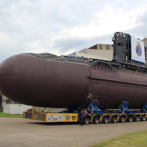 Imagem da galeria de imagem da página de Defesa. A imagem mostra o casco de um submarino sendo transportado por um caminhão de dentro da fábrica para a parte externa.