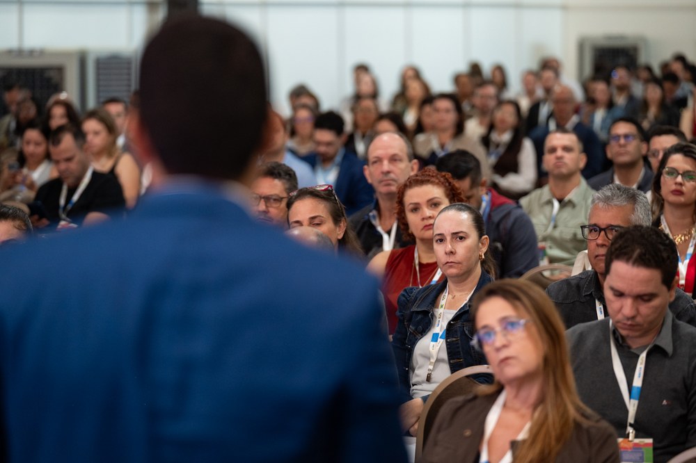 Foto com palestrante de costas, dando enfoque no publico da palestra com várias pessoas.