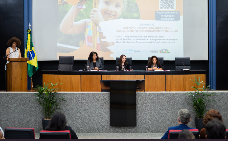Imagem de palco ao fundo com três mulheres em mesa e uma mulher à esquerda em púlpito