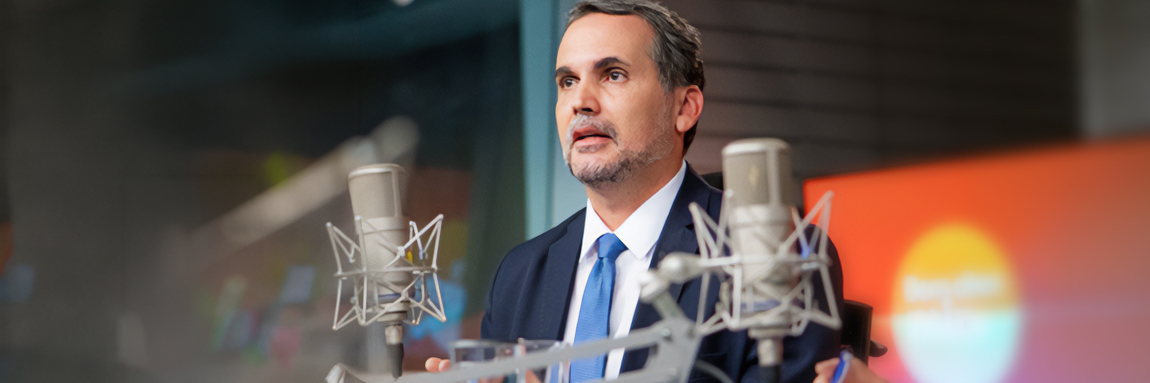 em primeiro plano microfone de rádio, ao fundo homem branco de terno concede entrevista