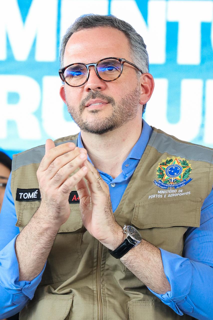 Homem de óculos e barba curta aparece em close, usando camisa azul e colete bege do Ministério de Po