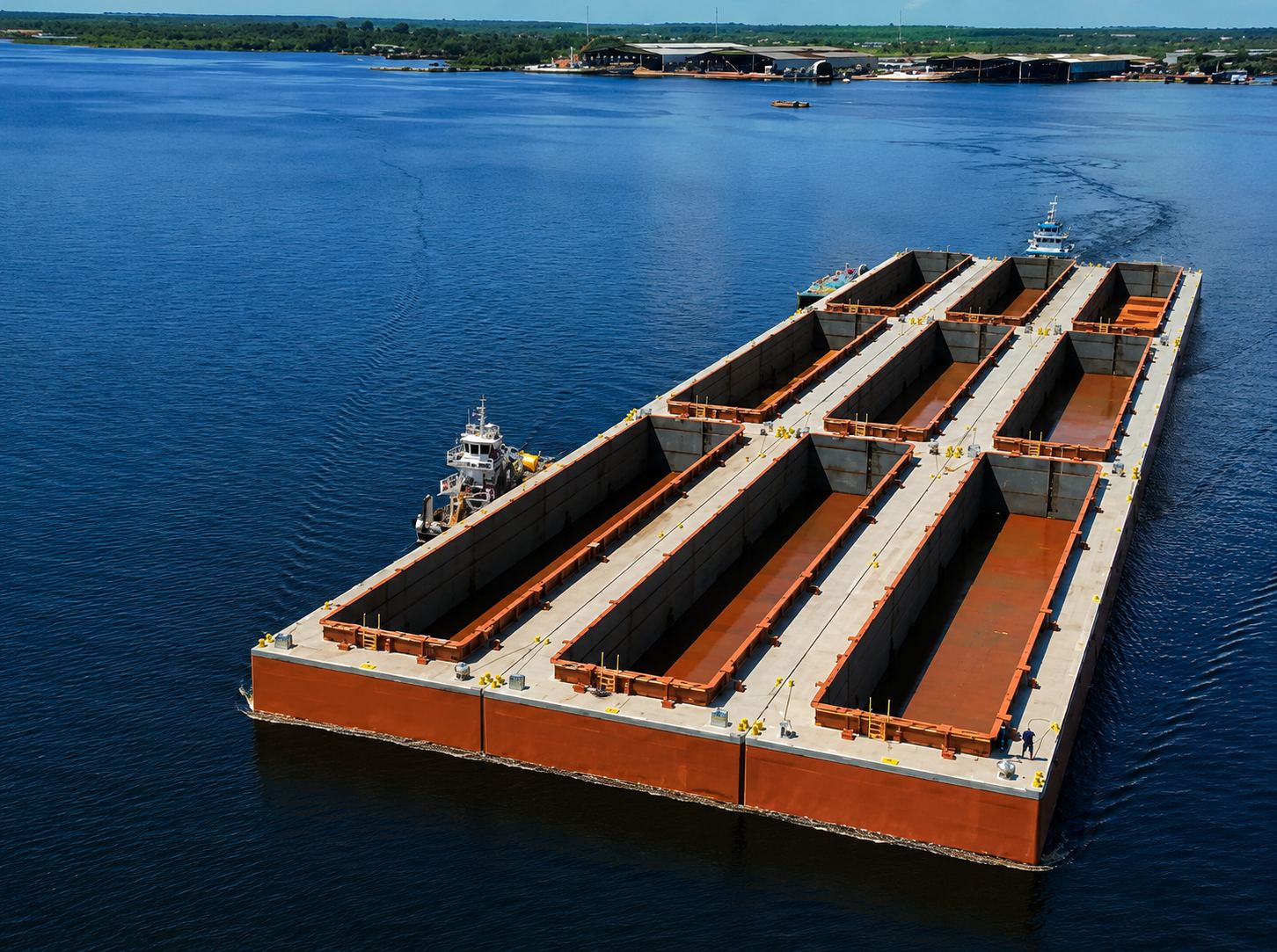 A imagem mostra uma balsa de carga navegando em um rio da Amazônia, conduzida por rebocadores. Ao fundo, aparecem instalações portuárias, destacando a importância das hidrovias para o transporte de cargas e o abastecimento da região Norte.