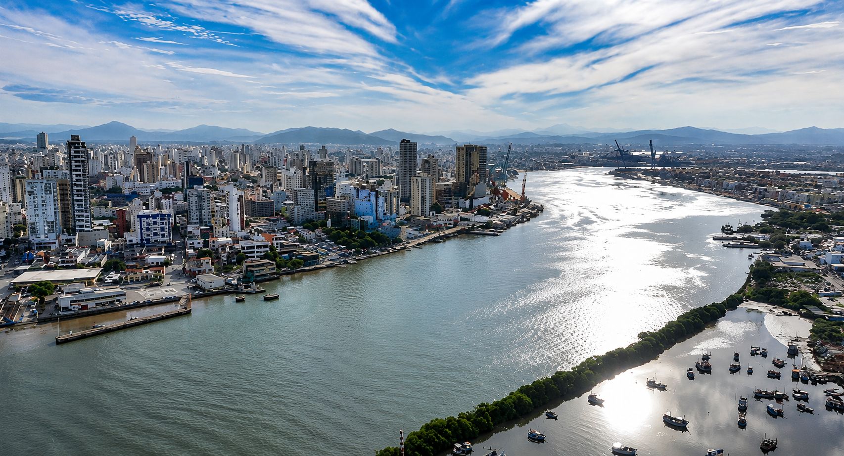 Vista aérea do canal de acesso ao Porto de Itajaí, em Santa Catarina. A imagem mostra um amplo trecho navegável que conecta o oceano às instalações portuárias e à área urbana da cidade. Nas margens do canal, destacam-se edifícios residenciais e comerciais, além de estruturas portuárias e guindastes ao fundo.
