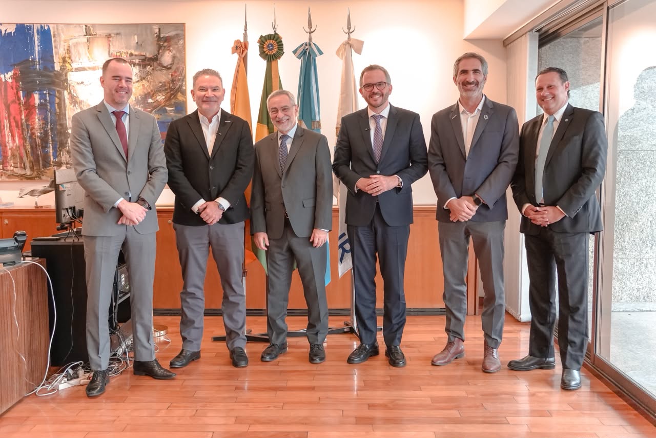 Seis homens de terno posam lado a lado em uma sala de escritório, sobre piso de madeira, entre eles está o ministro de Portos e Aeroportos, Tomé Franca. Ao fundo, há bandeiras em mastros e um quadro abstrato na parede. À direita, uma ampla janela de vidro deixa entrar a luz natural. Todos sorriem para a foto.