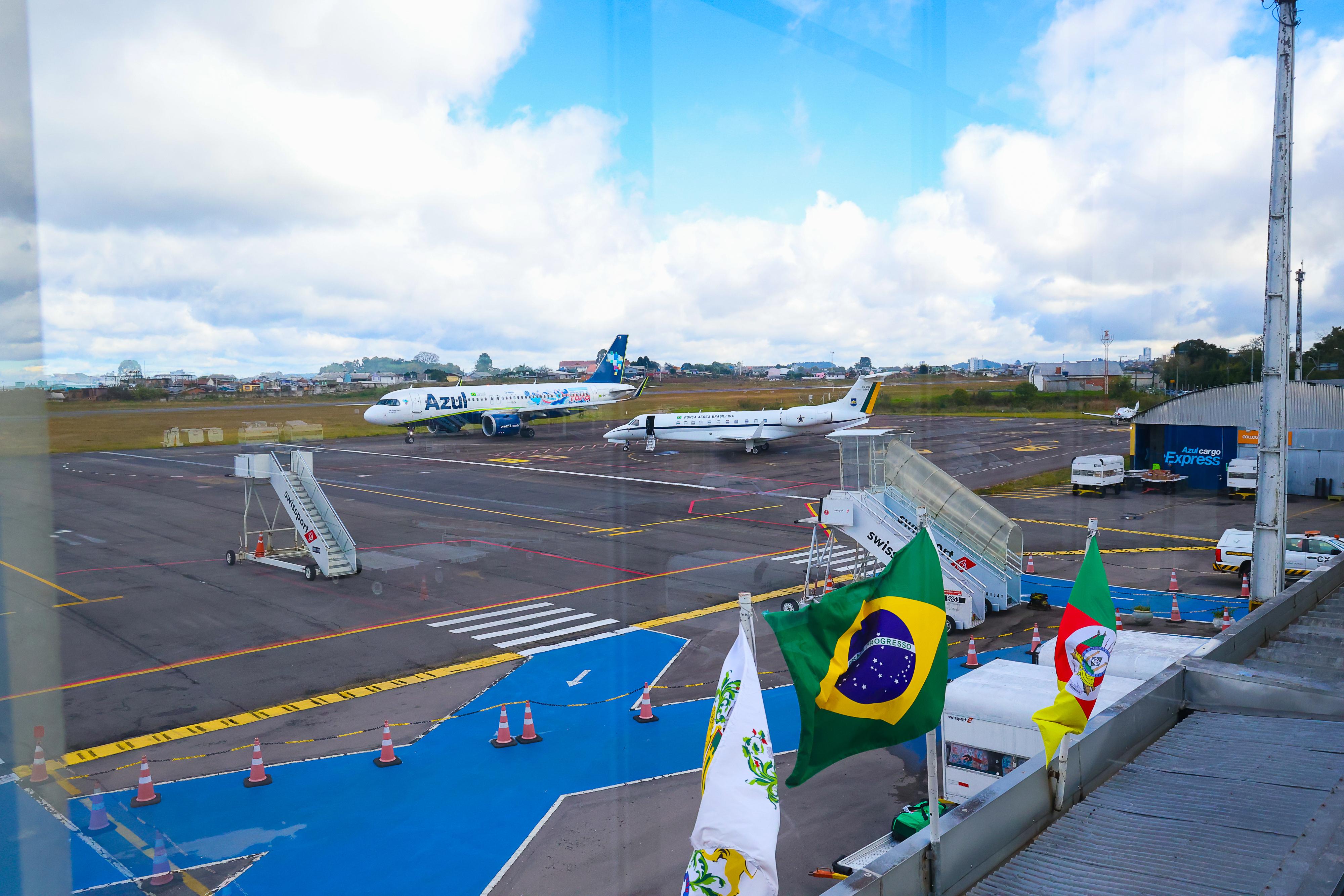 Foto mostra o pátio do aeroporto com dois aviões ao fundo. No canto direito estão estaiadas as band