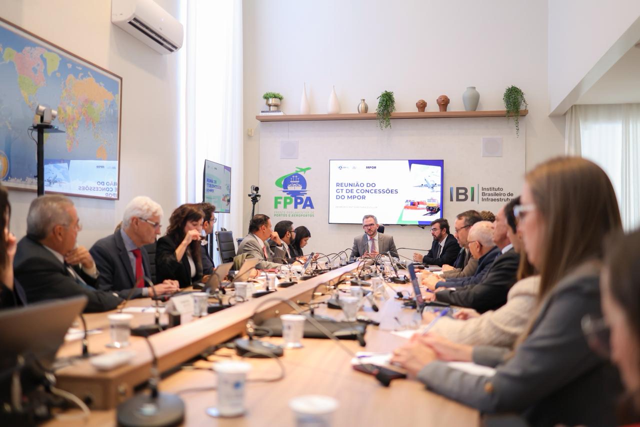 A foto registra uma reunião corporativa ou governamental de caráter formal, realizada em uma sala de conferências ampla, bem iluminada e de teto alto. Um grupo de aproximadamente 15 a 20 pessoas está reunido em torno de uma grande mesa de reuniões oval. O foco principal está ao fundo, onde um homem preside a reunião diante de uma tela de apresentação.