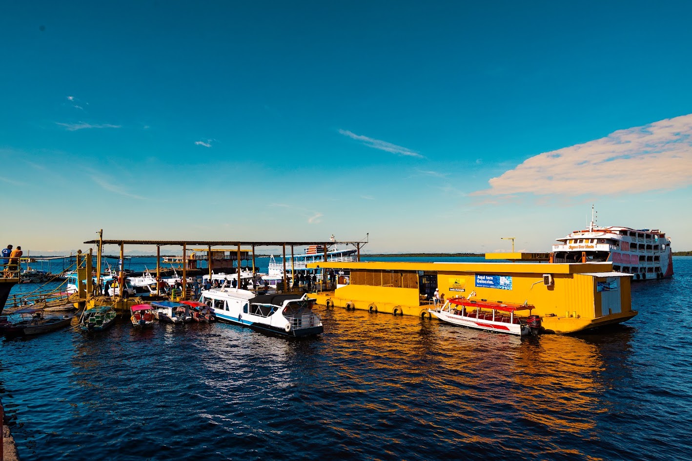 Fotografia do Porto da Manaus Moderna, em Manaus (AM), mostrando uma instalação portuária flutuante com embarcações atracadas para transporte de passageiros. Em destaque, um terminal amarelo sobre as águas do Rio Negro, cercado por barcos de pequeno porte e um navio regional ao fundo. O céu azul e a iluminação natural reforçam a paisagem típica da Amazônia.