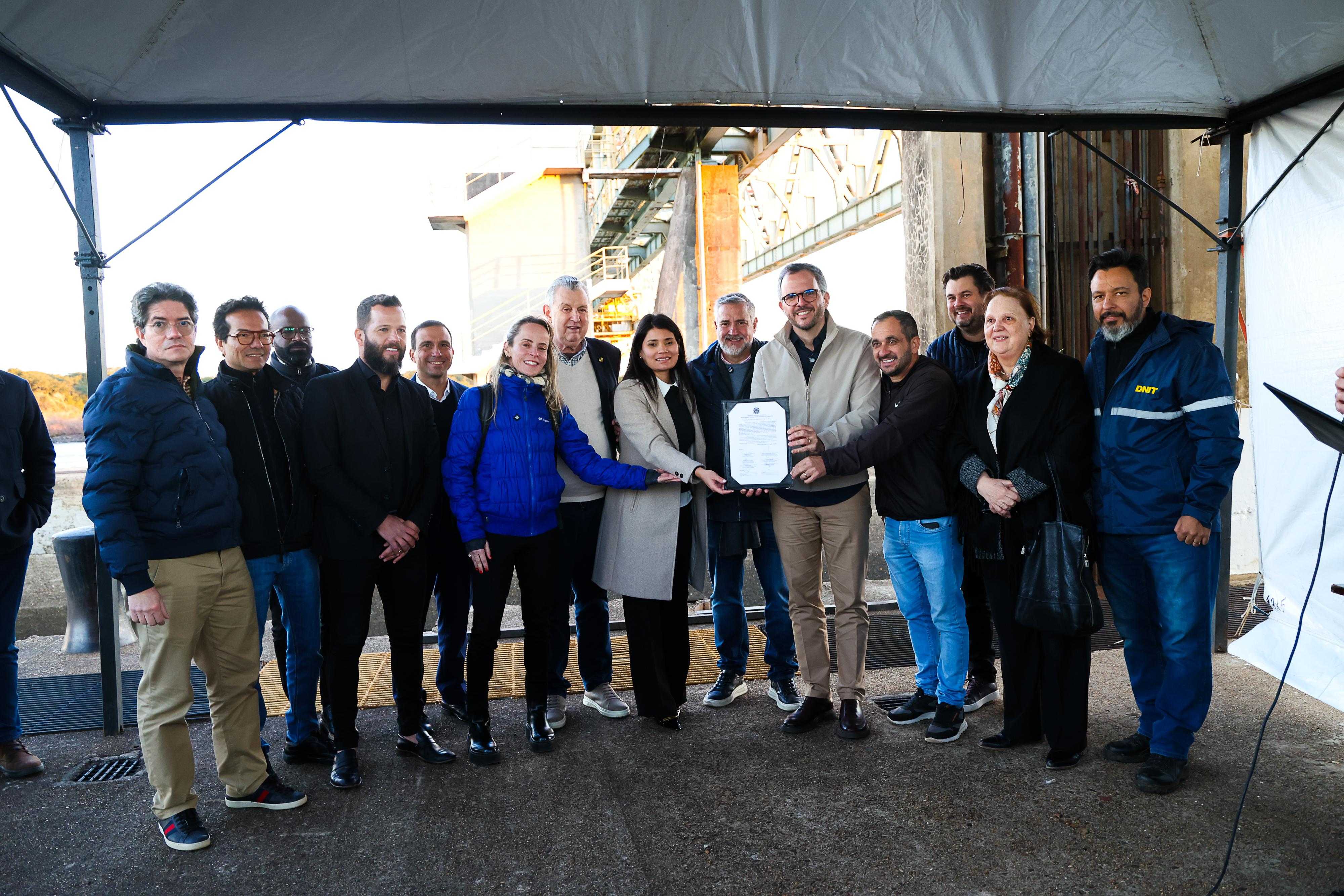 A imagem mostra 13 pessoas reunidas embaixo de uma tenda, pousando para uma foto oficial. No centro do grupo, o ministro de Portos e Aeroportos, Tomé Franca, segura um documento oficial emoldurado com capa escura. O ambiente ao fundo sugere uma área de infraestrutura portuária, com grandes estruturas de concreto e metal sob a luz do dia.